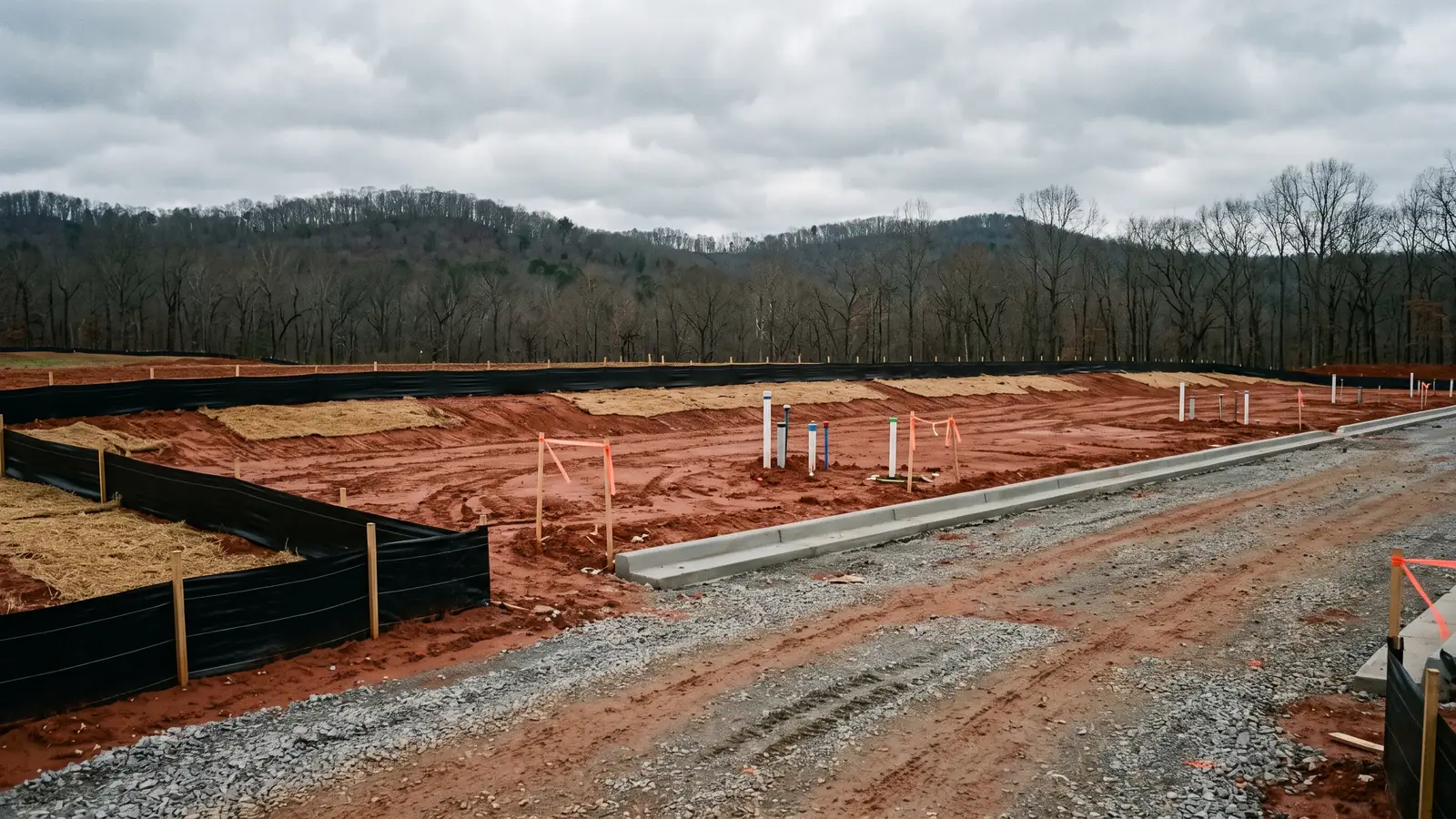 Early-stage commercial development site: a graded red-clay pad ringed with silt fencing and erosion-control matting, stubbed utility risers, fresh concrete kerbing and a gravel construction entrance, with wooded ridges behind.