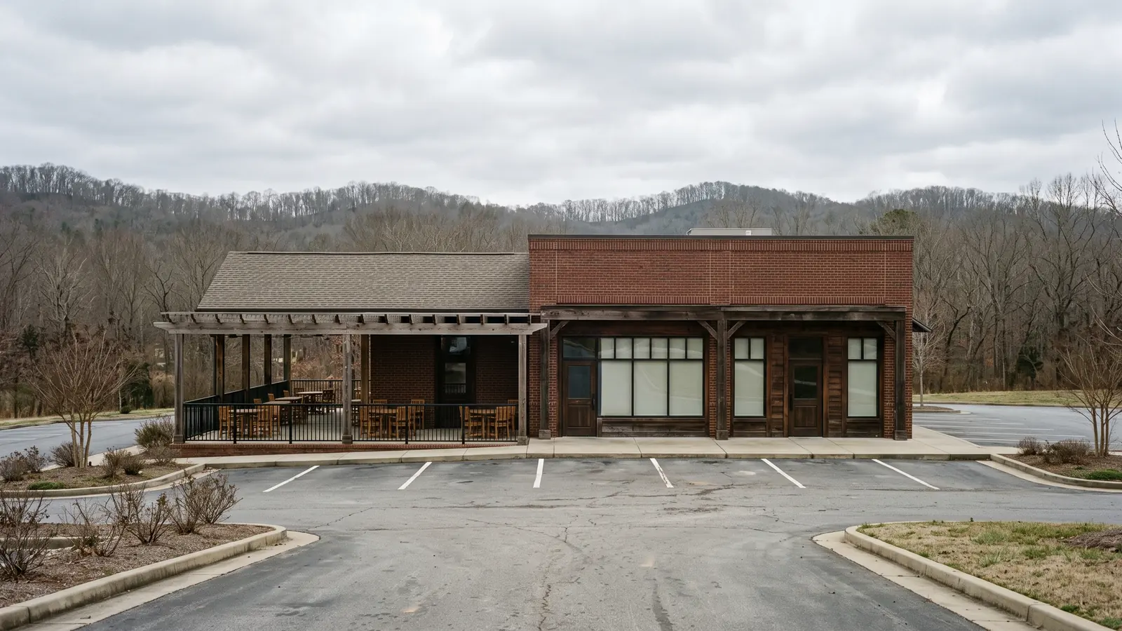 Freestanding restaurant building faced in brick and dark timber with a covered patio under bare pergola beams, black steel railings, large storefront windows and an empty striped parking lot in front.