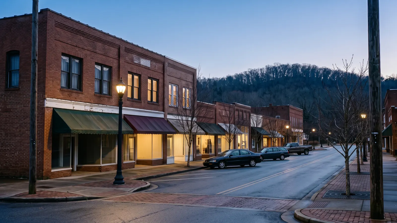 Street-level corner view of attached two-storey brick storefronts with canvas awnings, a brick paver sidewalk, a cast-iron street lamp, angled kerbside parking and bare street trees.