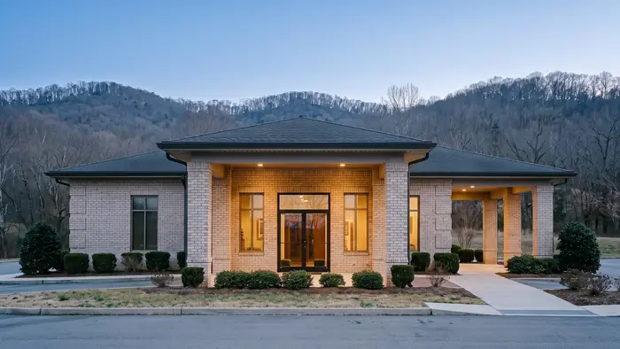 Single-storey light brick medical office building with a columned portico entry, tall bronze-framed windows, a covered drop-off lane and low clipped shrubs, wooded ridges in the background.