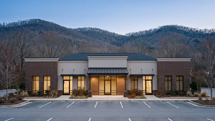 Single-storey brick and stucco professional office building with bronze storefront glazing and a covered entry canopy, an empty striped parking lot in front and bare winter hardwood ridges rising behind.