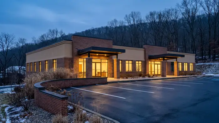 Single-storey brick and stucco professional office building with bronze storefront glazing and a covered entry canopy, an empty striped parking lot in front and bare winter hardwood ridges rising behind.