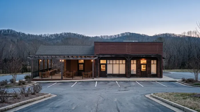 Freestanding restaurant building faced in brick and dark timber with a covered patio under bare pergola beams, black steel railings, large storefront windows and an empty striped parking lot in front.