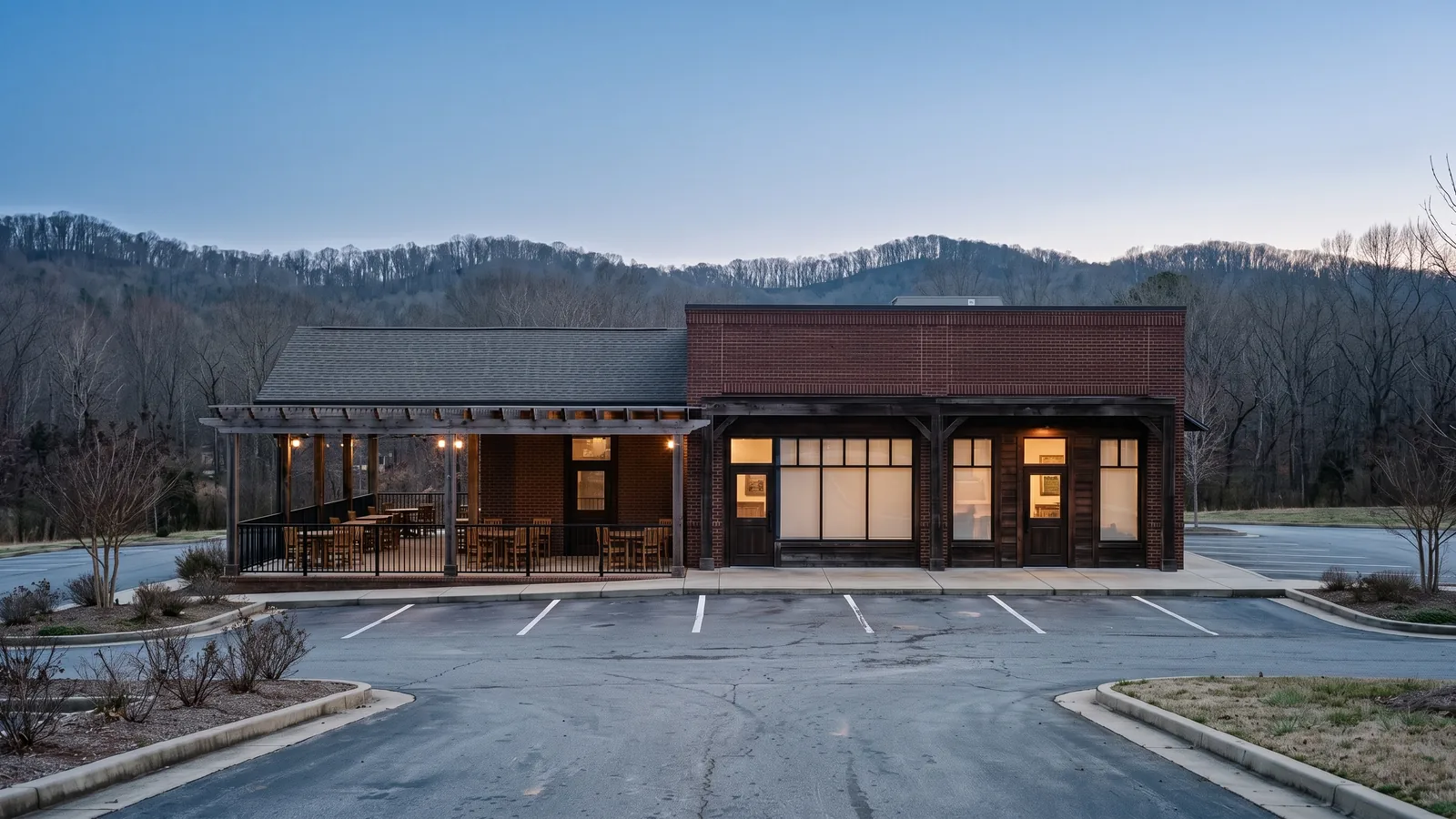 Freestanding restaurant building faced in brick and dark timber with a covered patio under bare pergola beams, black steel railings, large storefront windows and an empty striped parking lot in front.