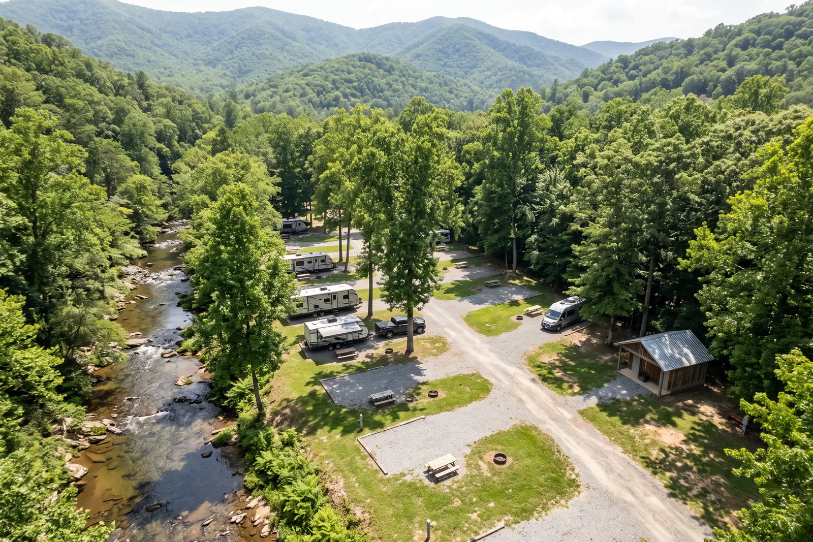 Illustrative image: an aerial view of a creekside campground with gravel RV pads, travel trailers, picnic areas and a small bathhouse under tall trees, mountains behind.
