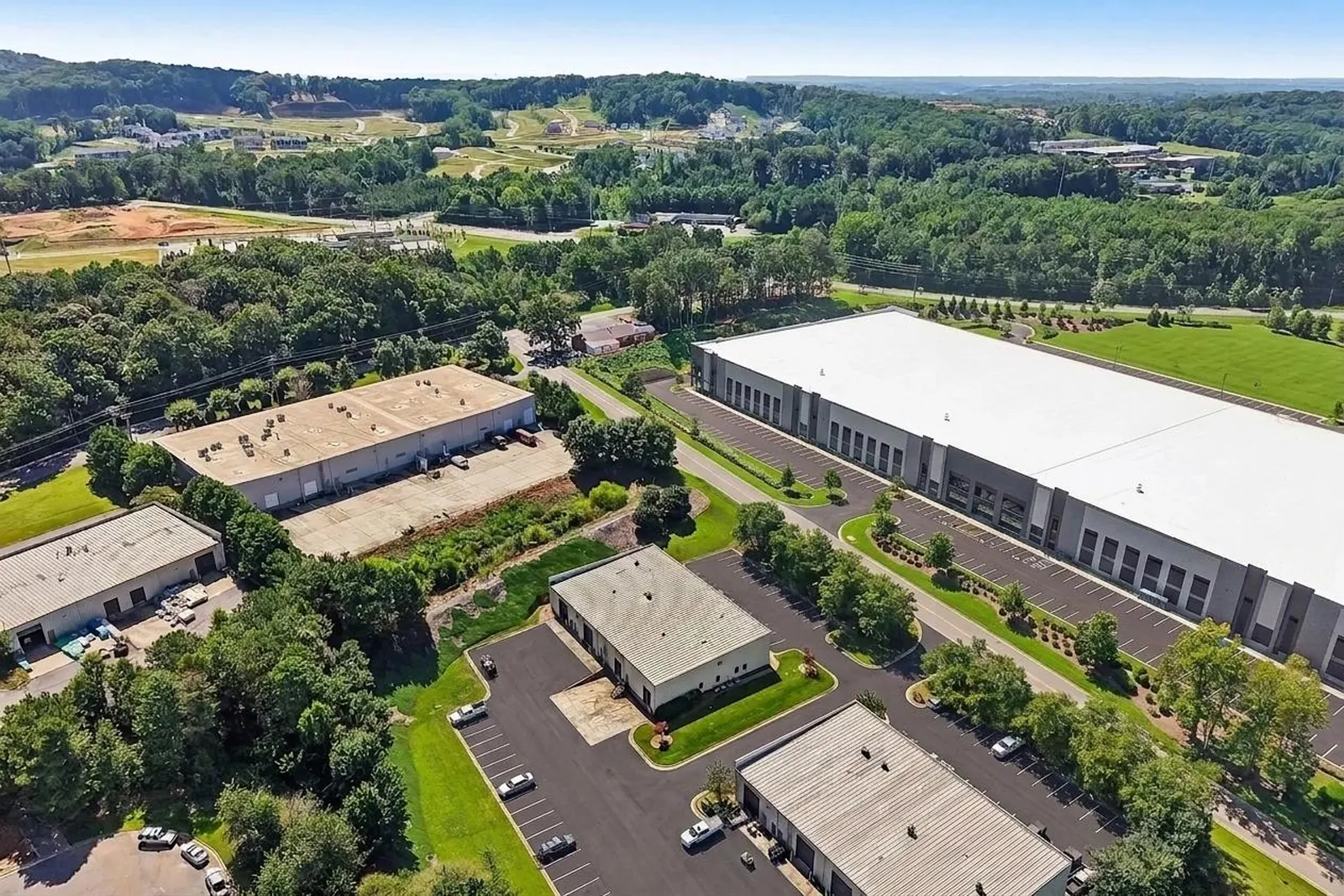 An aerial view of an industrial park with several flat-roofed office-warehouse buildings, parking lots, green lawns and wooded hills beyond.