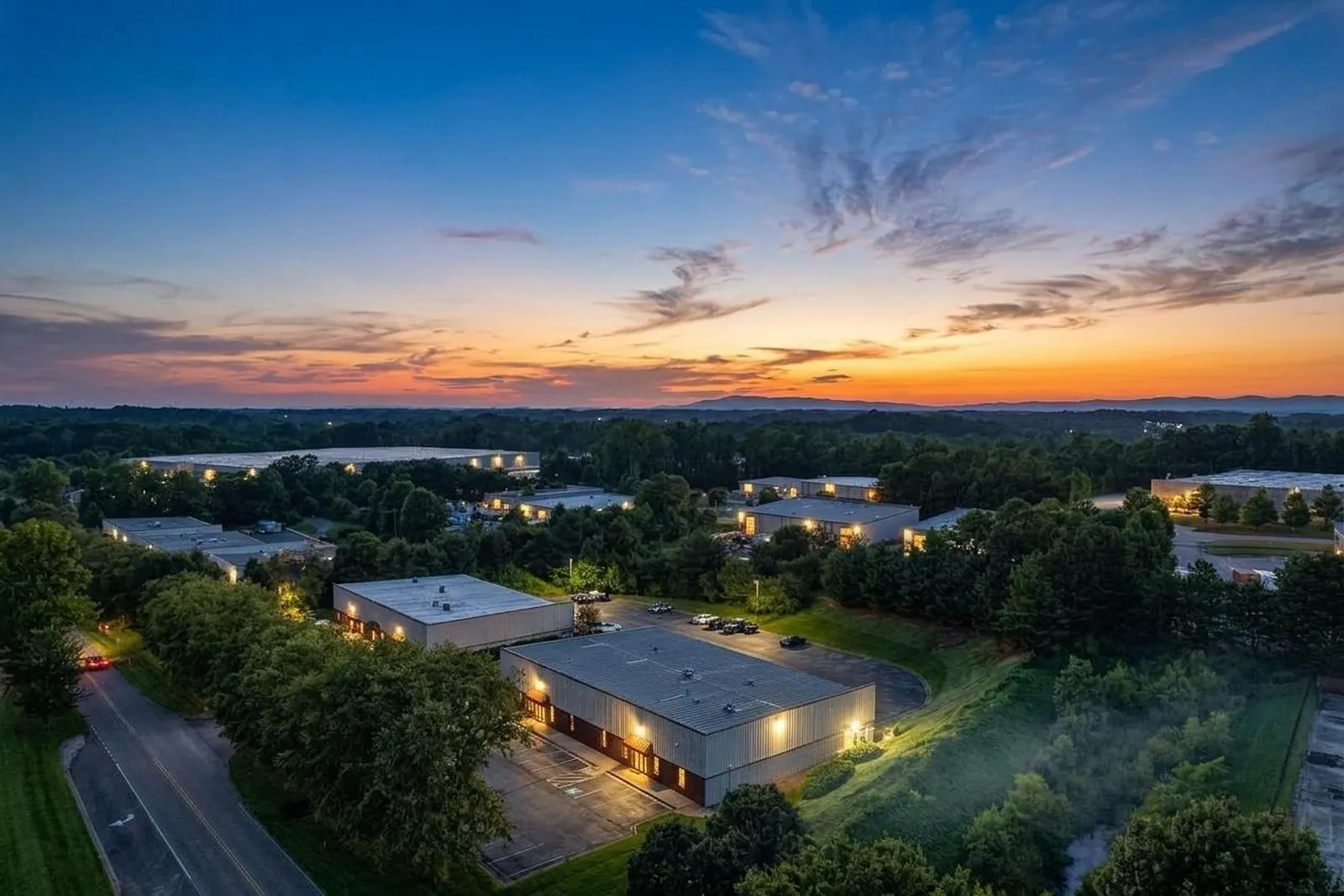 A dusk aerial view of a business park with lit office-warehouse buildings among trees, a winding road and a sunset sky over distant hills.