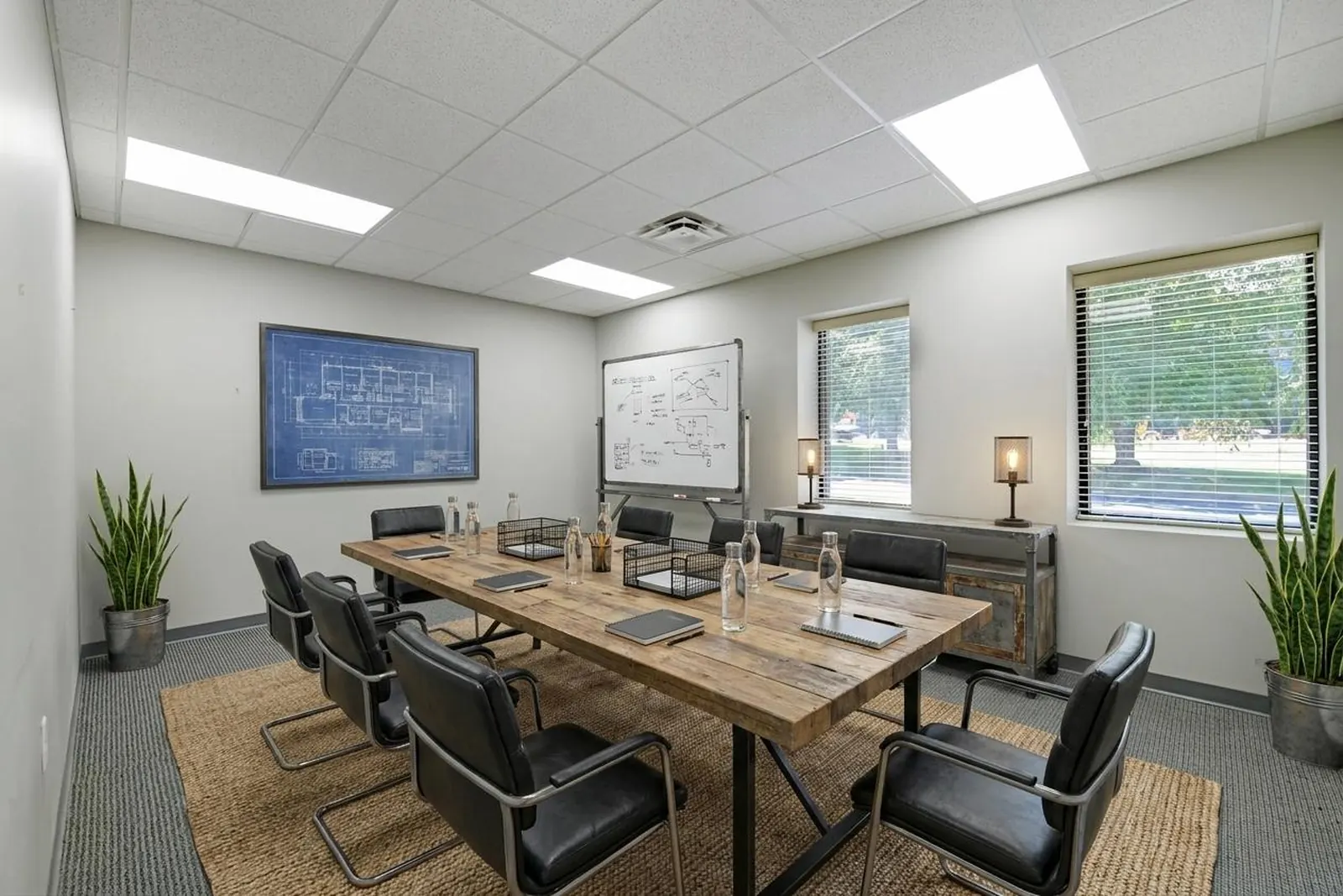 A virtually staged conference room with a long wood table, black chairs, a wall-mounted screen and framed art on pale walls.