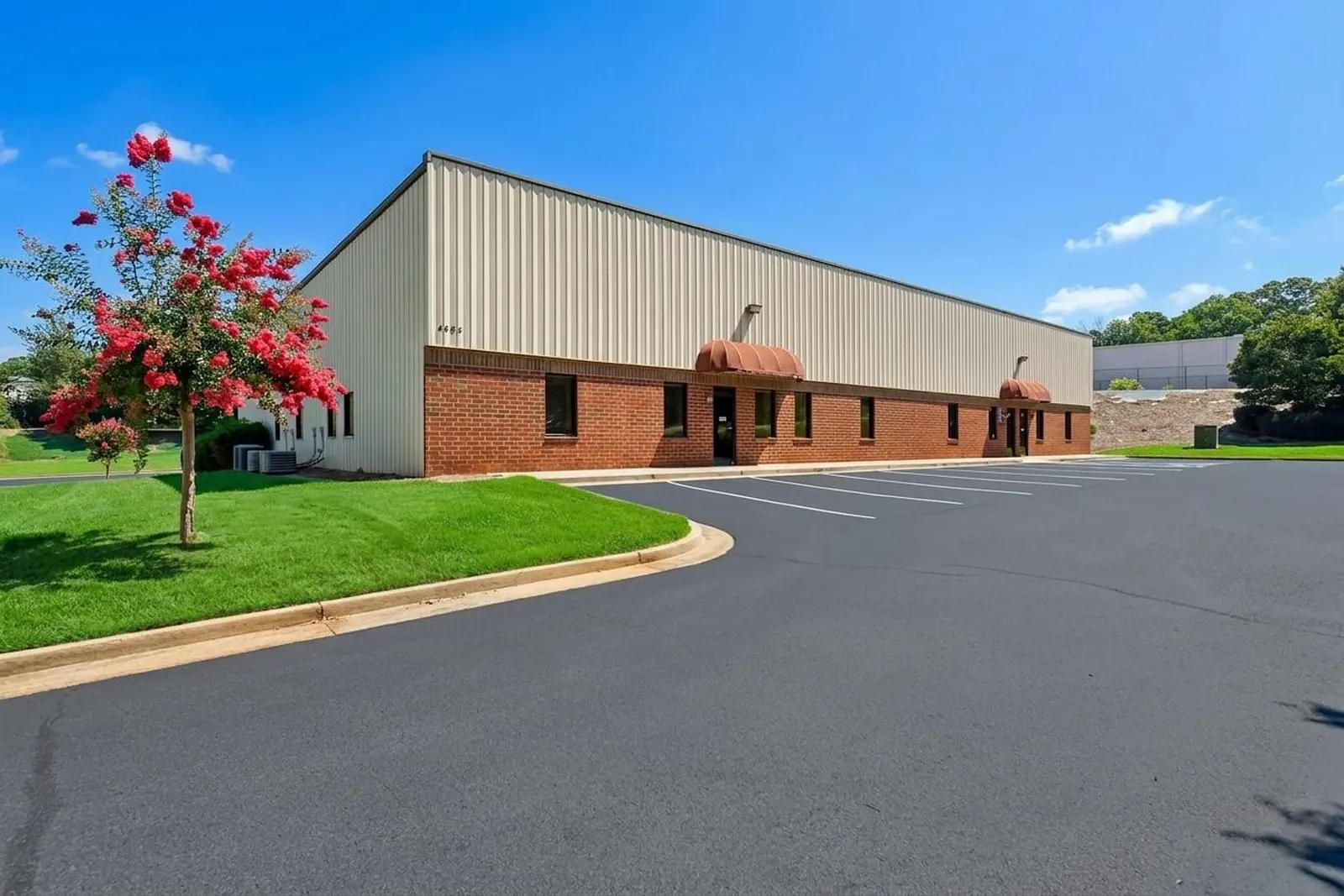 A long brick and metal office-warehouse building with red awnings over the entries, a blooming crape myrtle on a lawn island and a curving paved drive.