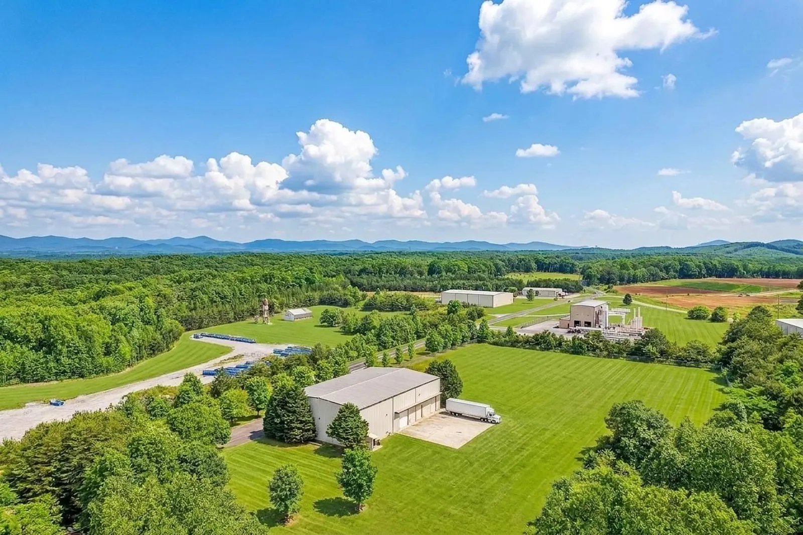 After: the same drone view with the lot shown as green lawn around the metal warehouse, forest and mountains beyond.