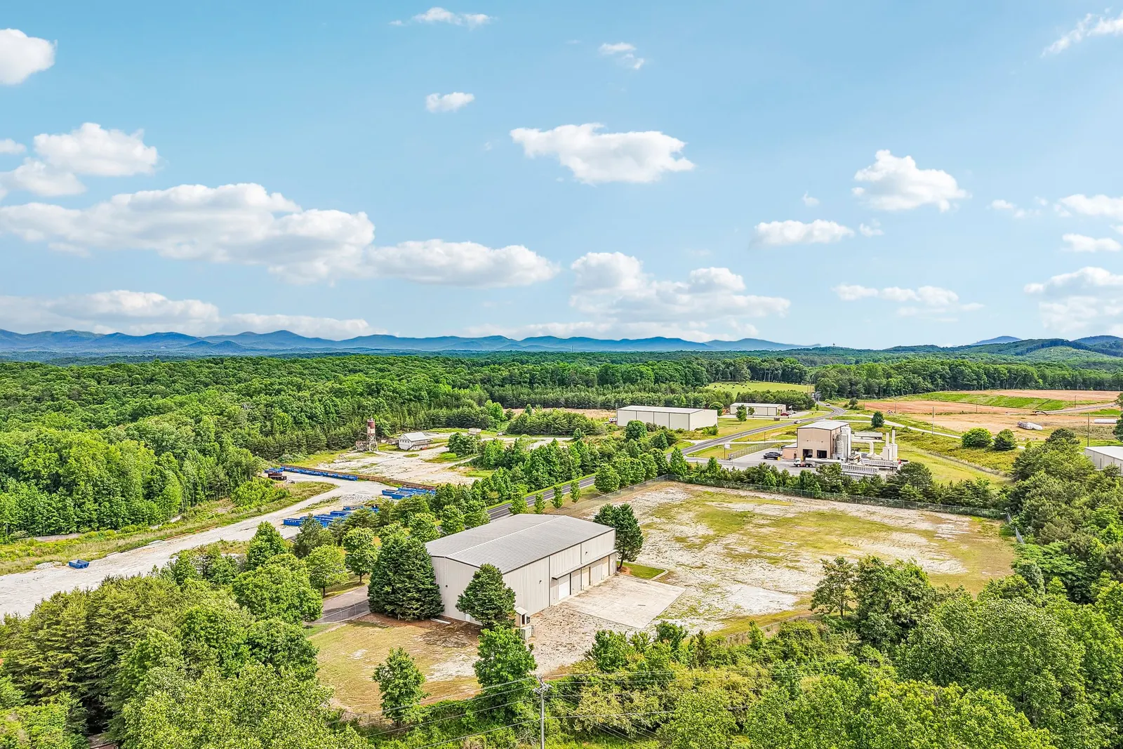 Before: a drone view over a metal warehouse on a bare gravel and brown-grass lot surrounded by forest, with mountains on the horizon.