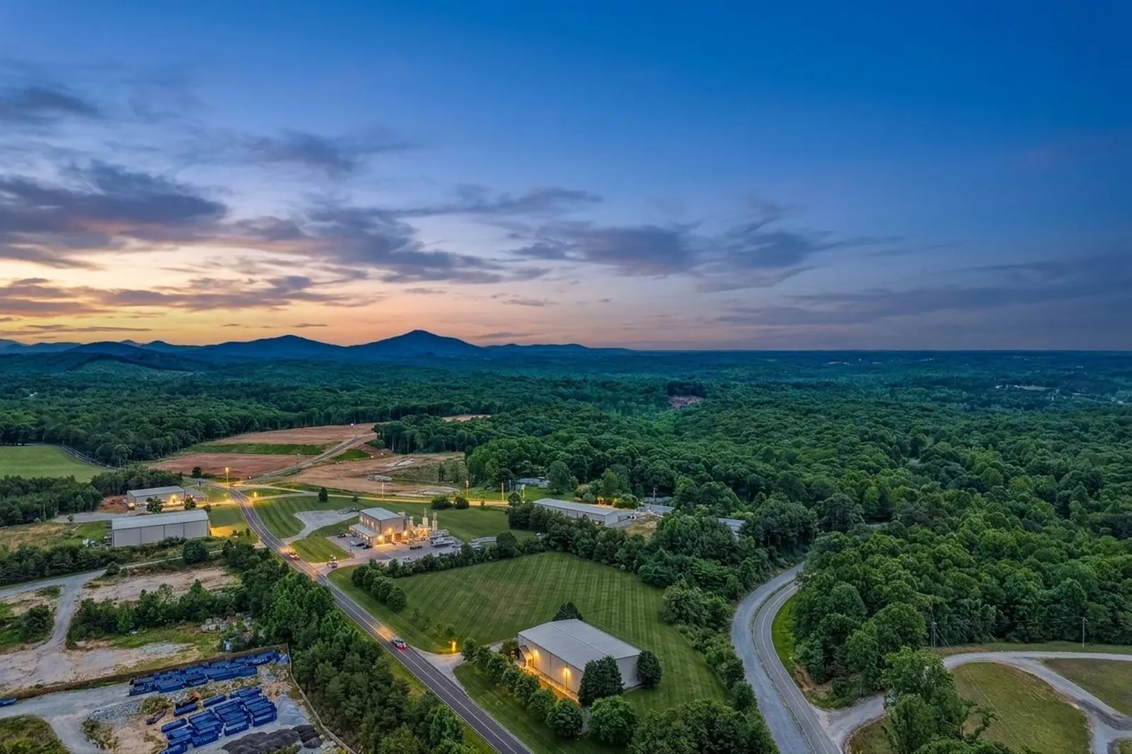 A dusk aerial view over a lit metal warehouse on a green lot beside a winding road, forested hills rolling to a line of blue mountains under a fading sky.