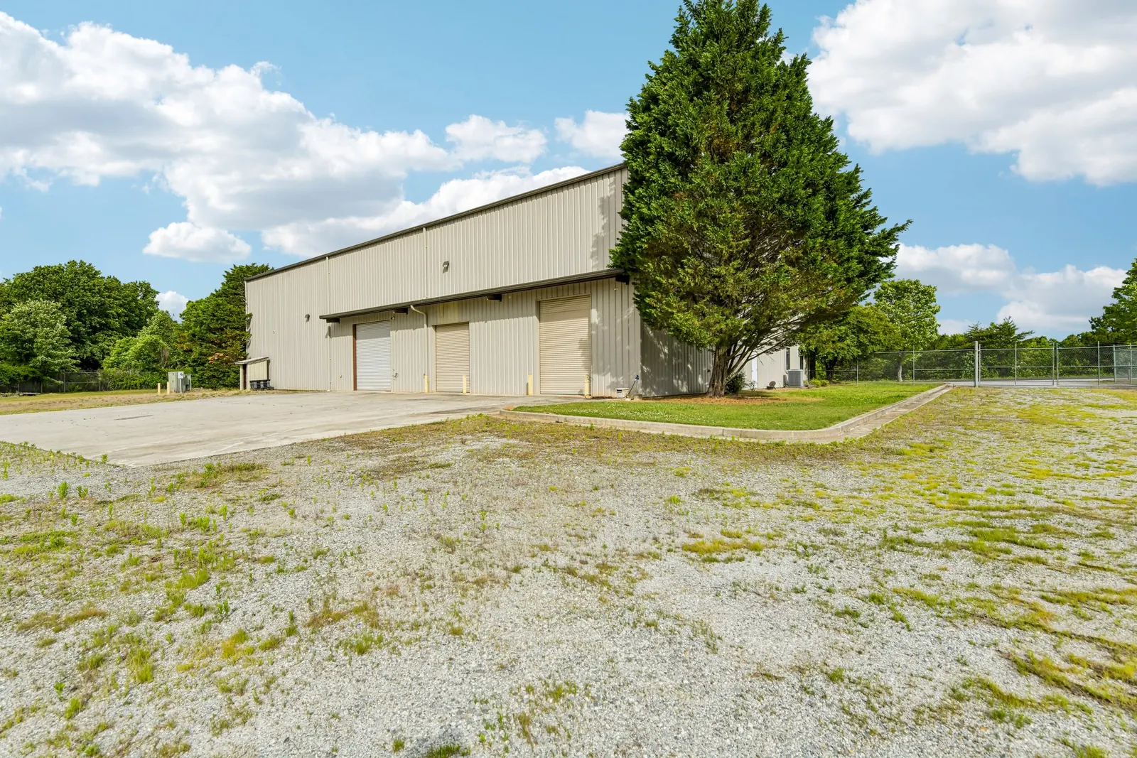 Before: the drive-in side of a tan metal warehouse with three overhead doors, a gravel yard and weedy, patchy ground in front.