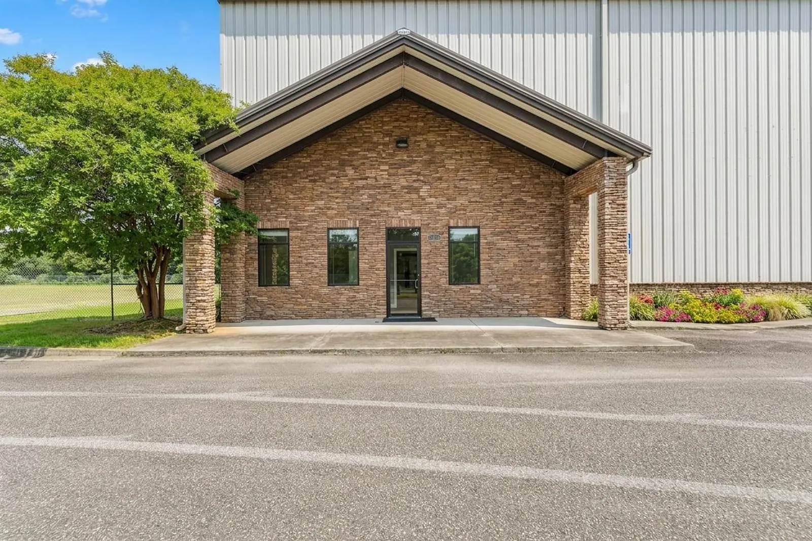 The stone and brick office wing of a metal industrial building, with a gabled roof, three windows and a glass entry door off a paved drive.