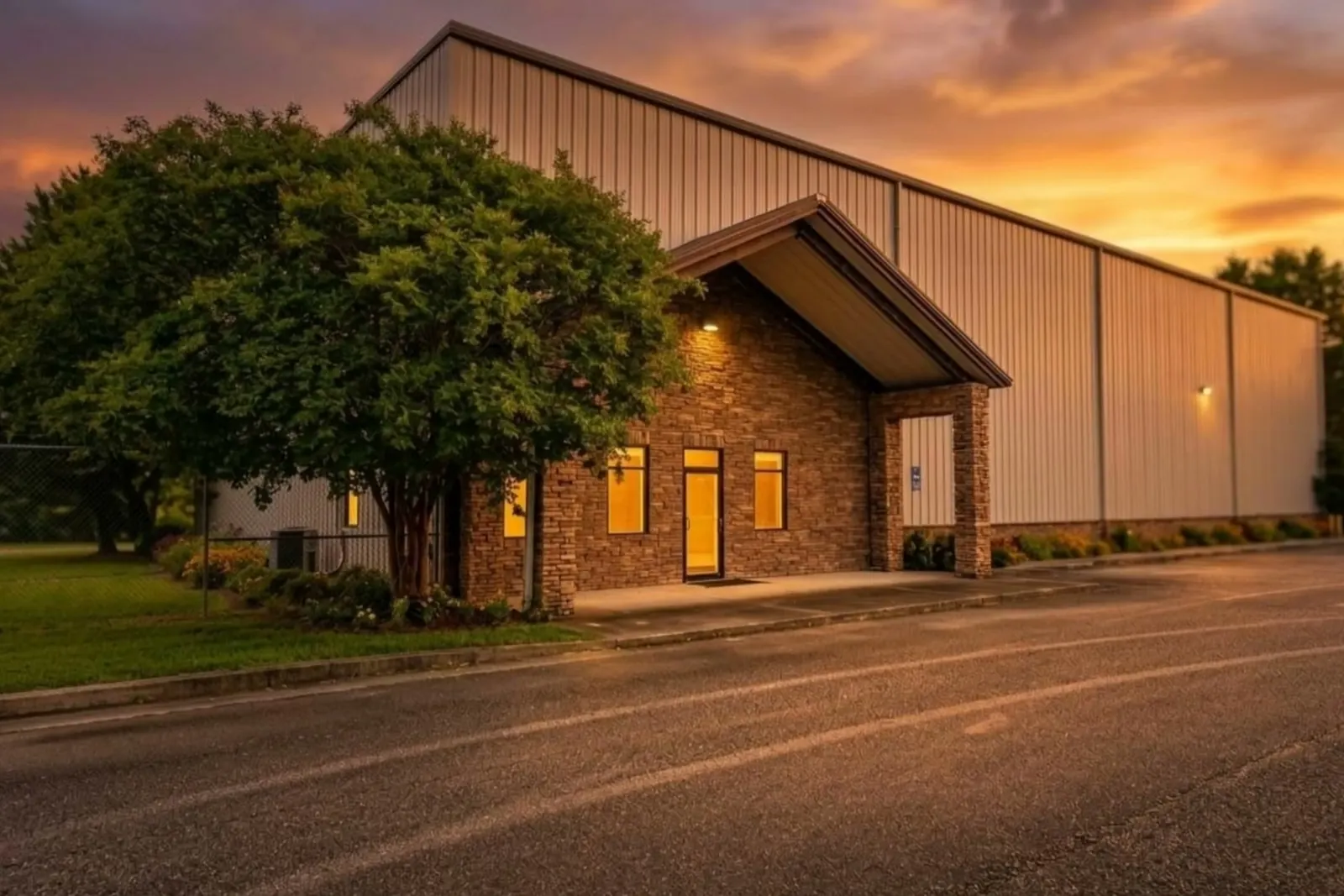 A metal industrial building with a stone and brick office entry at sunset, lights glowing at the entrance and windows, a large shade tree in front, a paved lot and an orange sky behind.