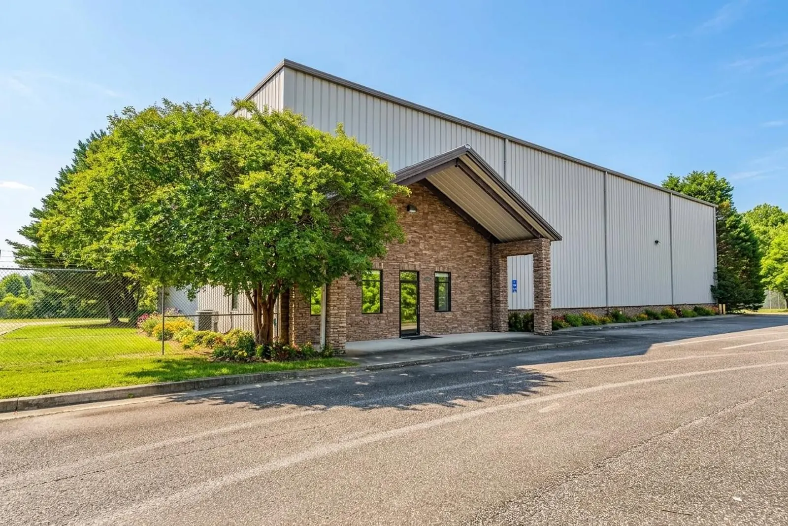 A metal industrial building with a brick and stone office entry behind a large shade tree, a paved drive curving past a green lawn.