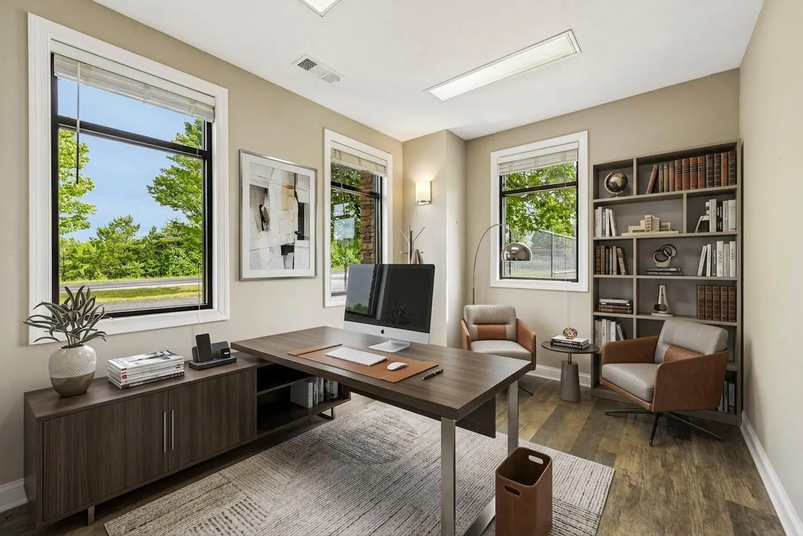 A virtually staged private office with a dark wood desk, a computer monitor, a lounge chair, a bookshelf and two windows looking out to trees.