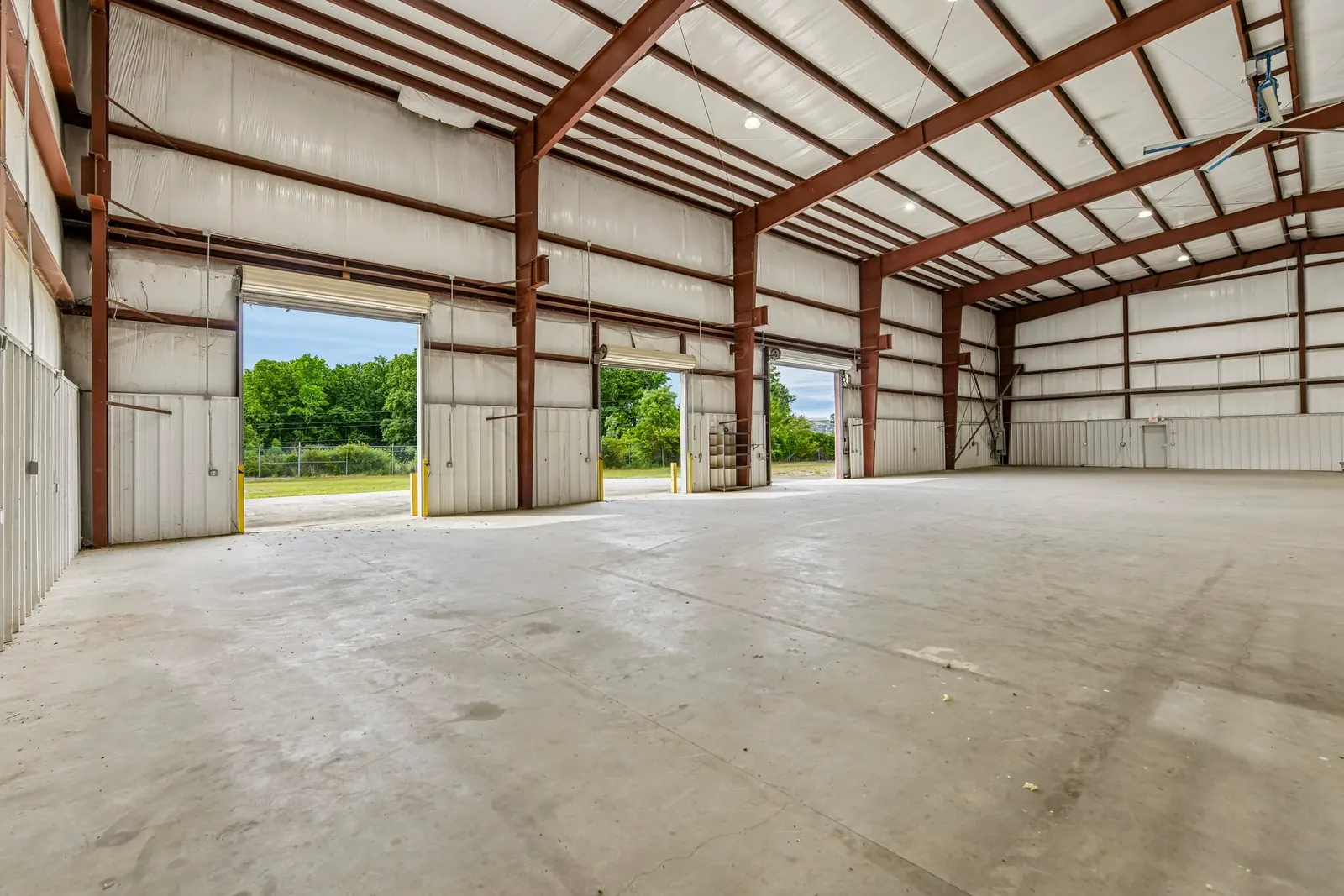 Before: an empty clear-span metal warehouse with red steel framing, a bare concrete floor and two open drive-in doors looking out to trees.