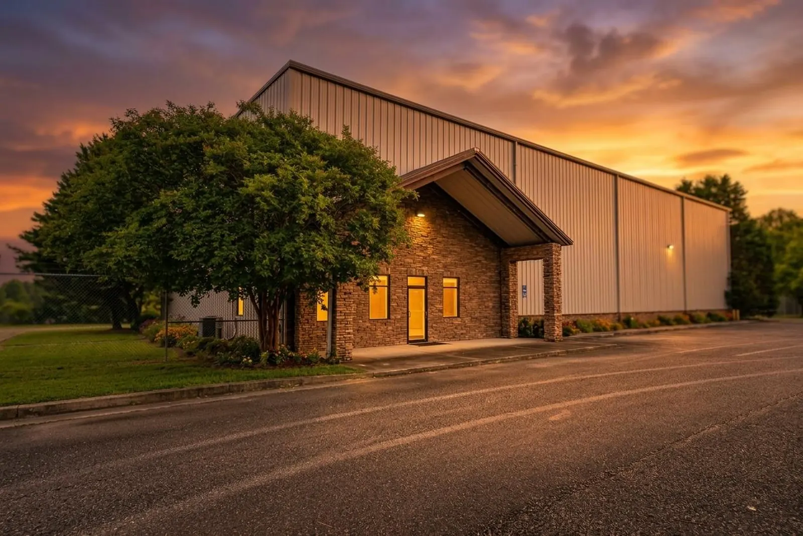 A metal industrial building with a stone and brick office entry at sunset, lights glowing at the entrance, a large shade tree in front and an orange sky behind.