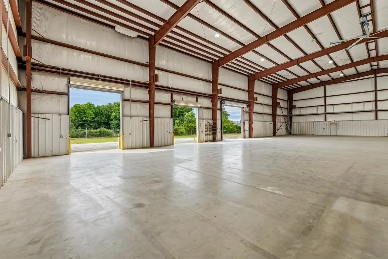 The inside of a clear-span metal warehouse with red steel framing, LED high-bay lights, a polished concrete floor and two open drive-in doors framing green trees outside.