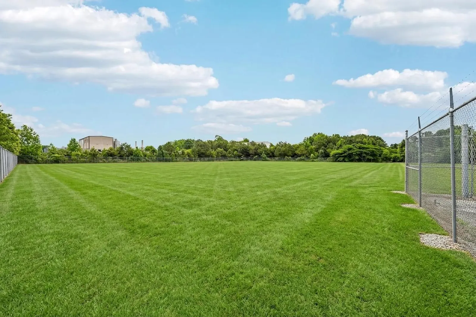 After: the same fenced industrial yard shown as a striped green lawn under a blue sky, with the chain-link fence on the right and the metal warehouse in the distance.