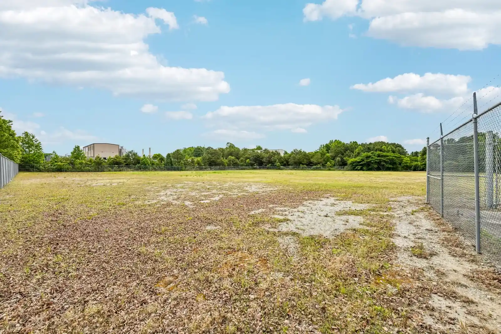 Before: a fenced industrial yard of patchy brown grass and bare gravel under a blue sky, a chain-link fence on the right and a metal warehouse far in the distance.