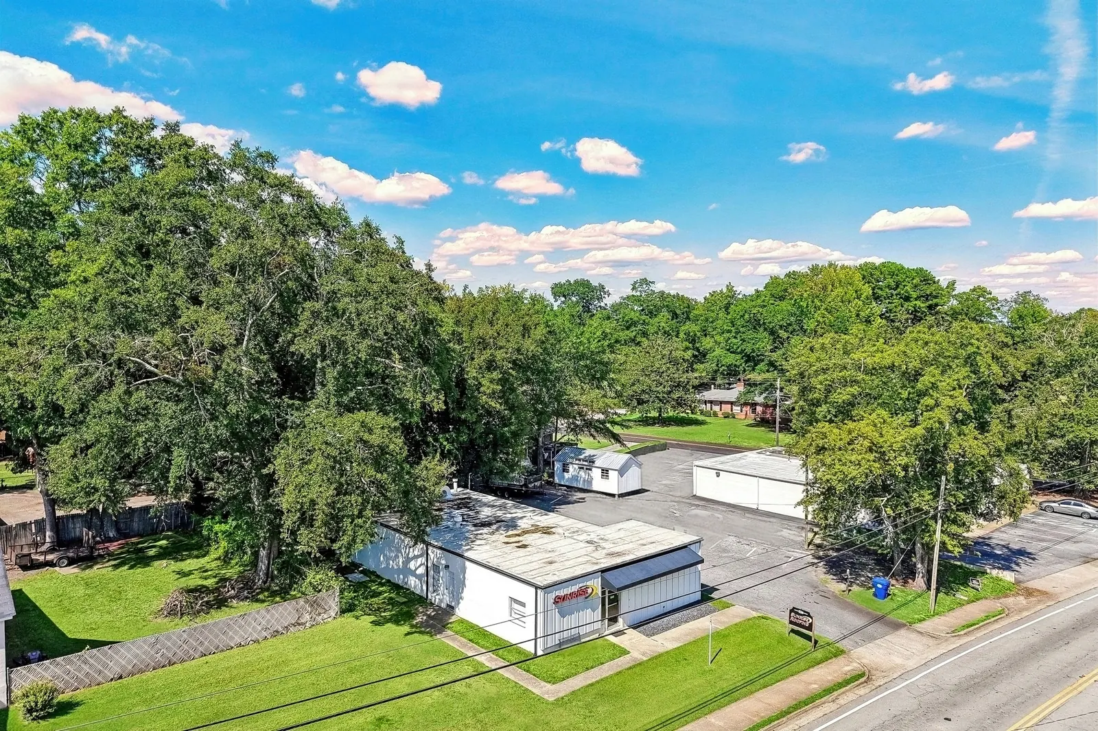 A drone view of a white flat-roofed commercial building with a paved lot and green lawn on a tree-lined street in a small town.