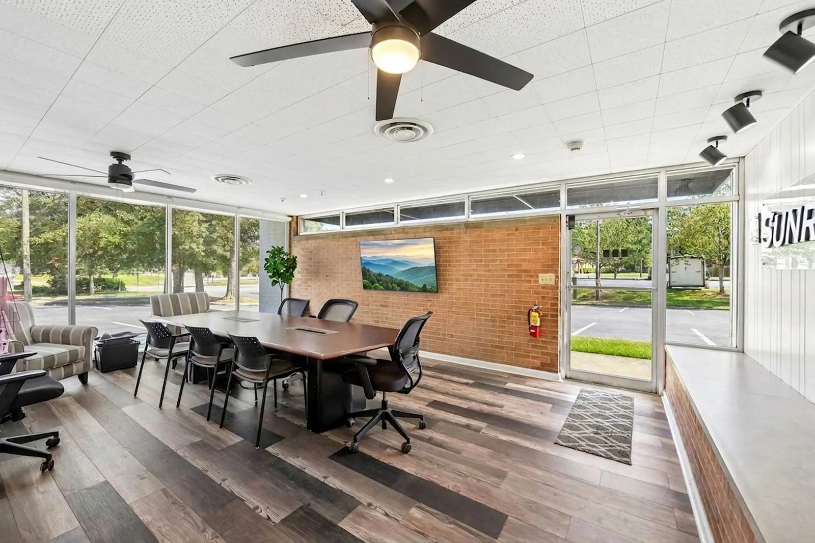 An open meeting area with a wood conference table, black chairs, a red brick wall with a wall-mounted screen and a wall of windows.