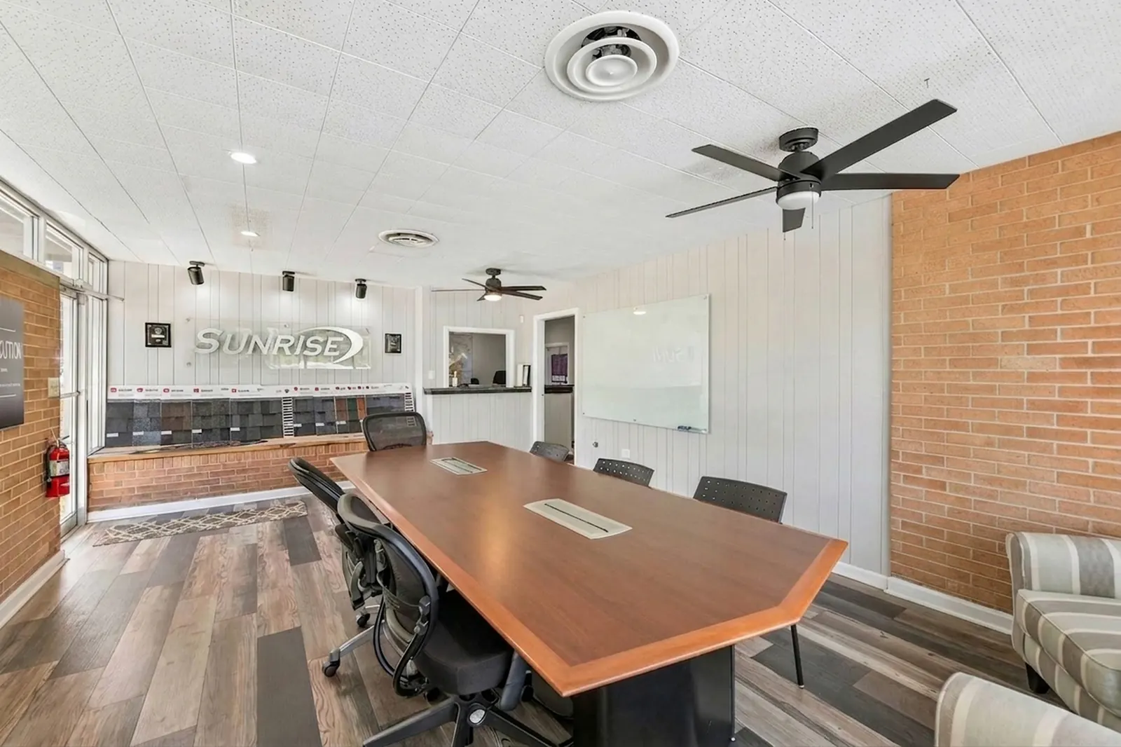 A conference room with a long wood table and black chairs, a red brick accent wall, ceiling fans and glass doors at the far end.