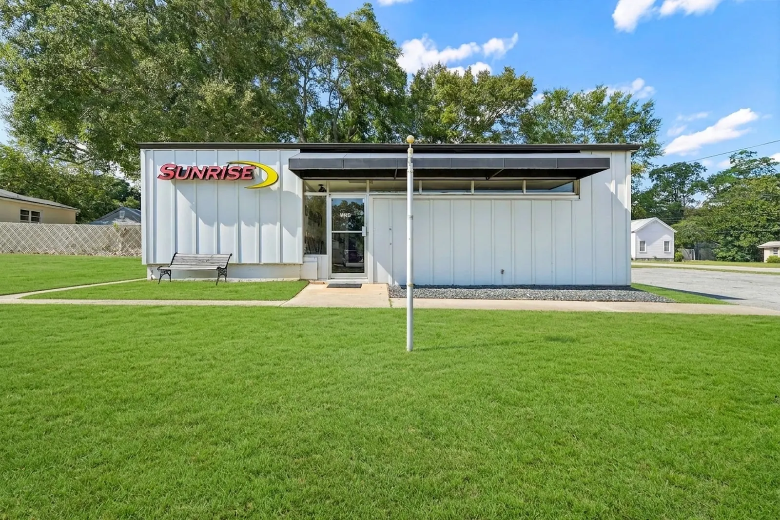 A white single-storey commercial office building with a dark roof overhang, a bench, a flagpole and a wide green front lawn under a blue sky.