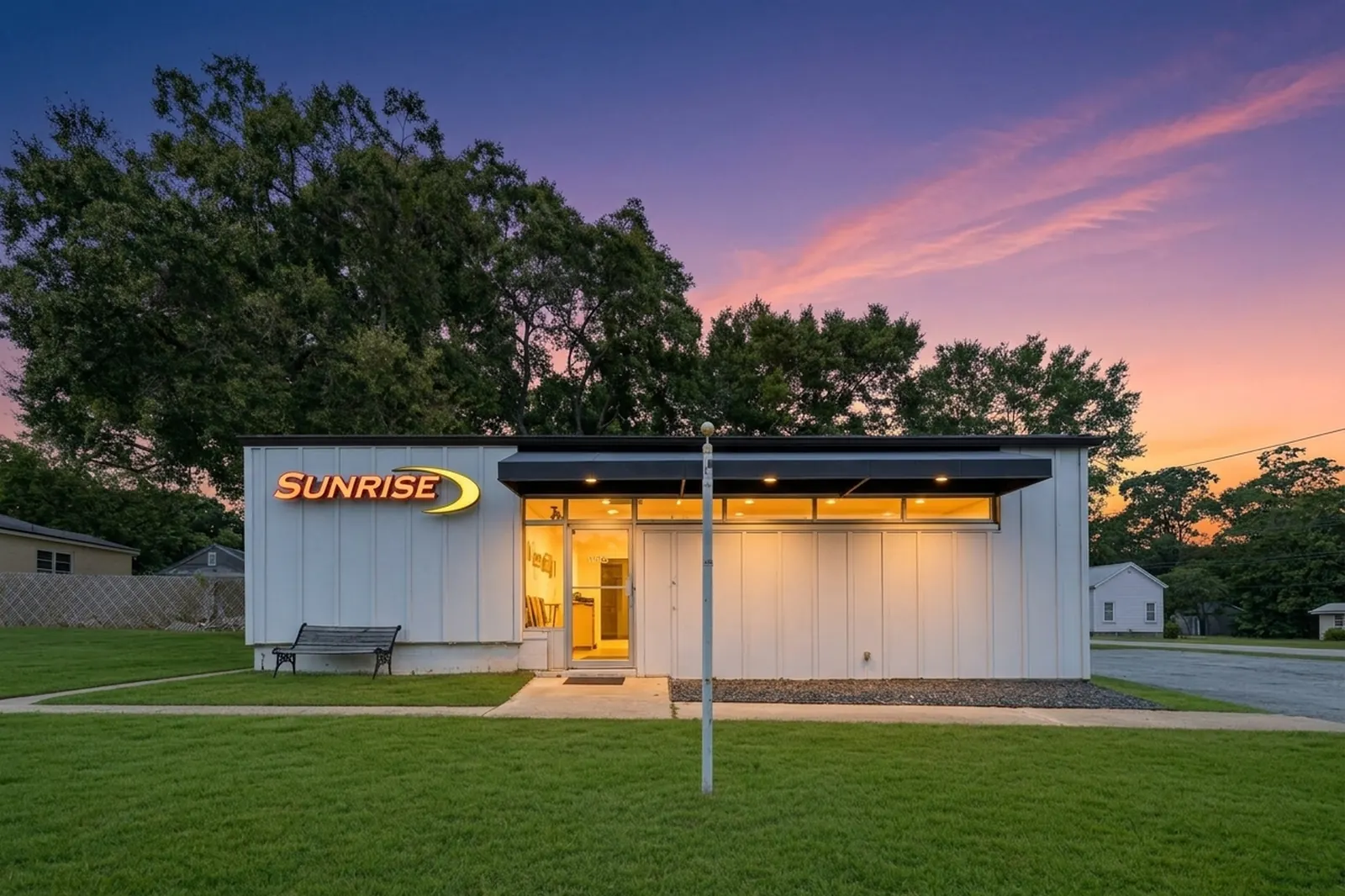 A white single-storey commercial office building at dusk with lit windows, a flagpole and a broad green front lawn under a pink and orange sky.
