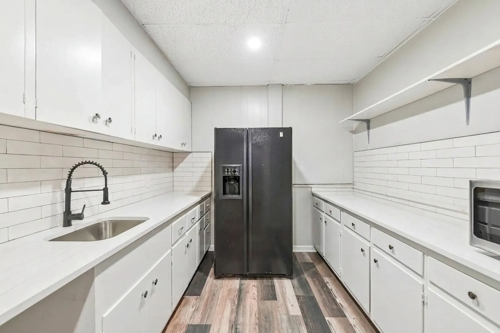 A small office kitchen with white cabinets, a white tile backsplash, a stainless sink and a black side-by-side refrigerator.