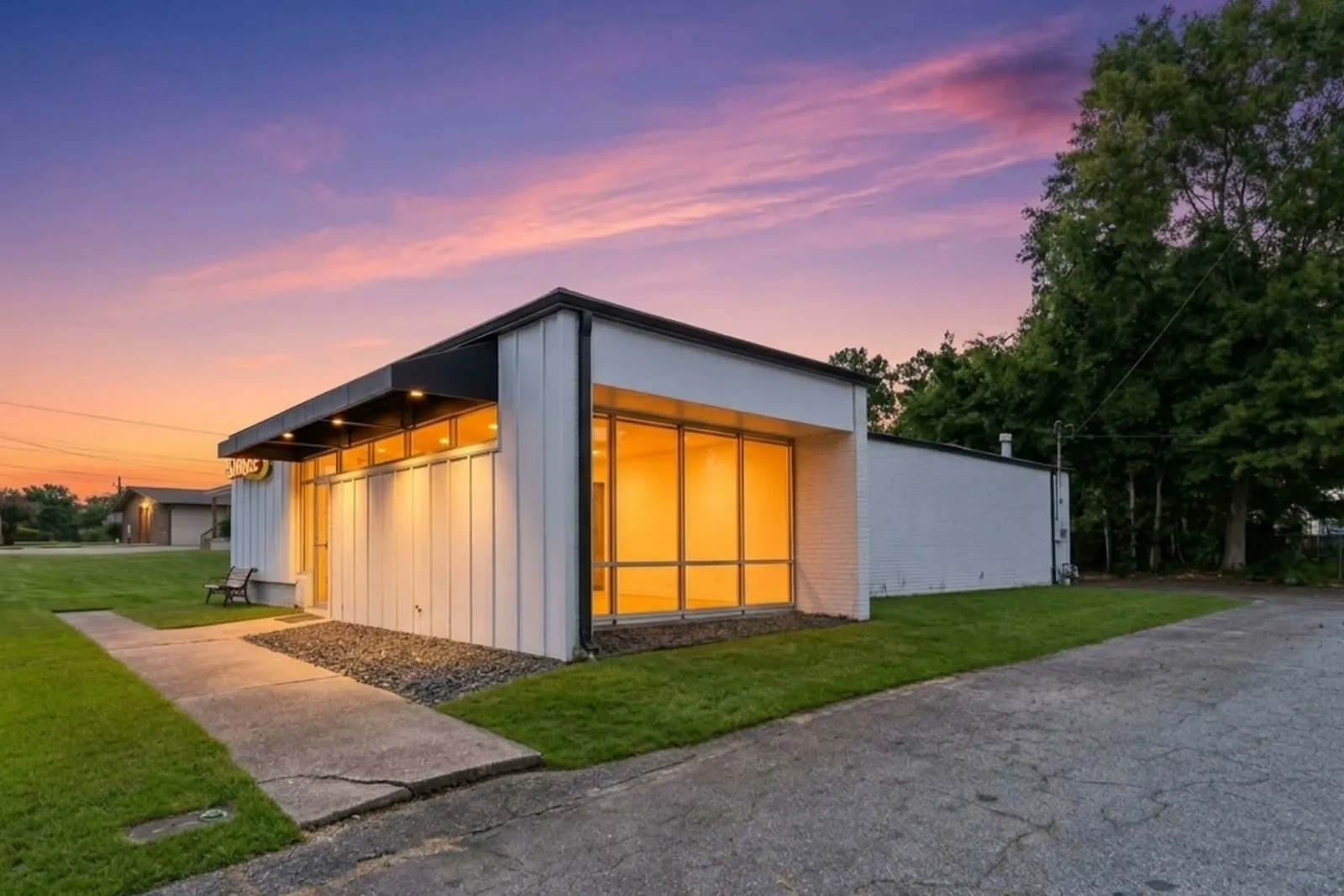 A white single-storey commercial office building at dusk with warm light glowing through tall front windows, a gravel border, a green lawn and a cracked asphalt drive, trees silhouetted against a pink and violet sky.