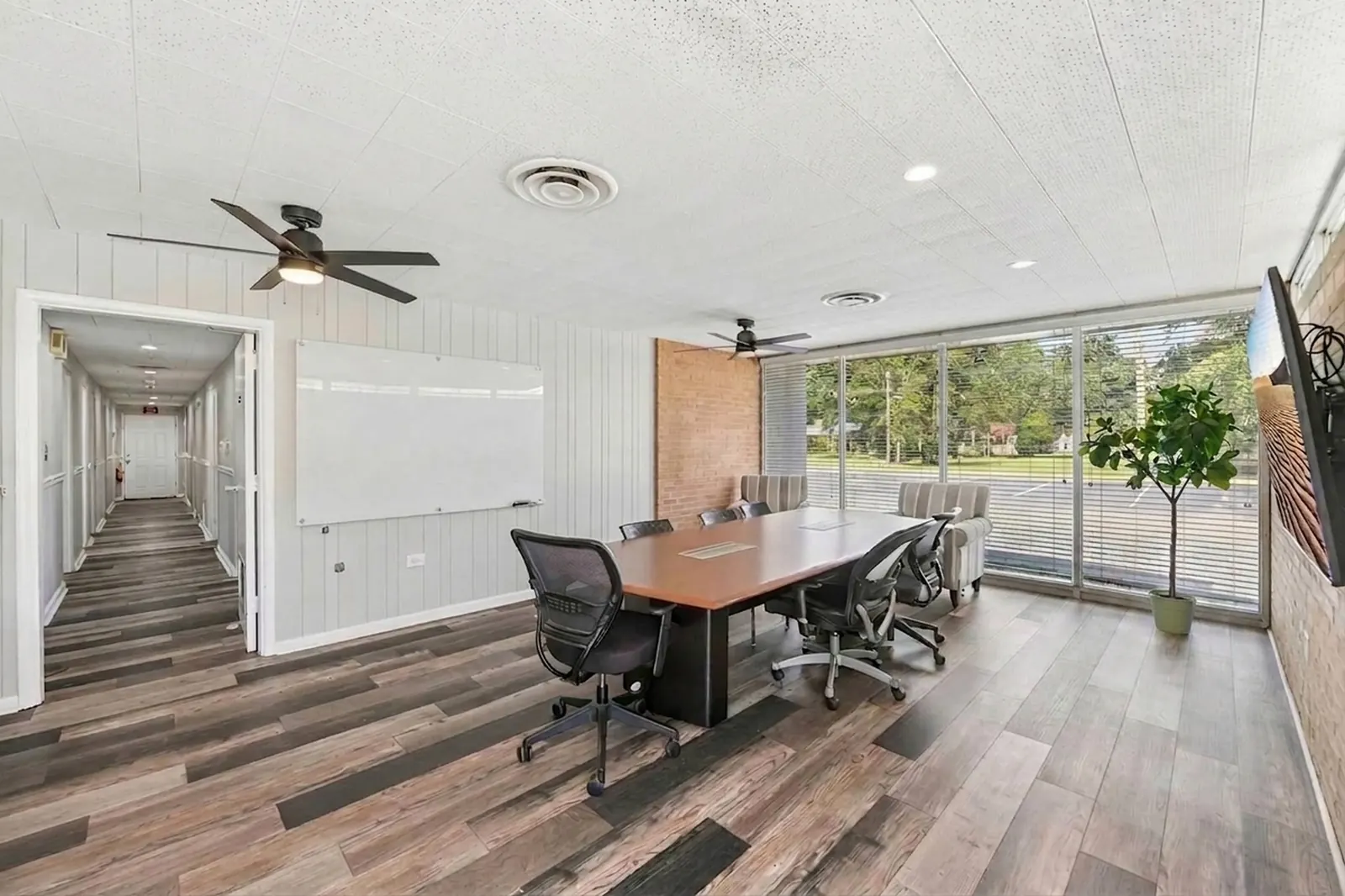 A bright workroom with a long table and office chairs, a whiteboard wall, a corridor leading back and floor-to-ceiling windows.