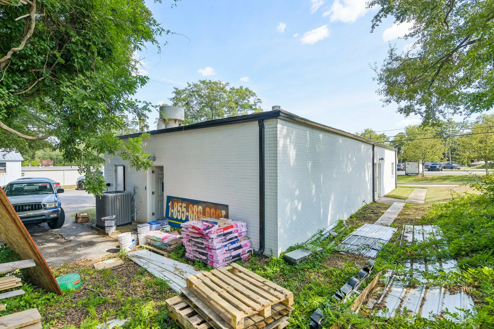 Before: the side of a white painted-brick commercial building with pallets, stacked roofing material, sheet metal and debris in an overgrown yard, a pickup parked at left.