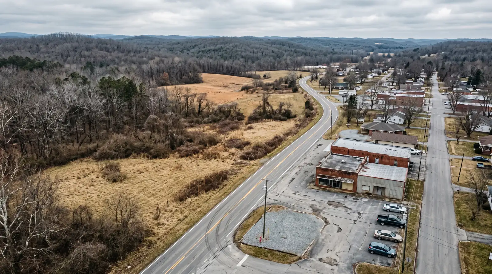 Aerial view of a two-lane road at the edge of a small town, with undeveloped grass and wooded parcels on one side and low commercial buildings and parking on the other.