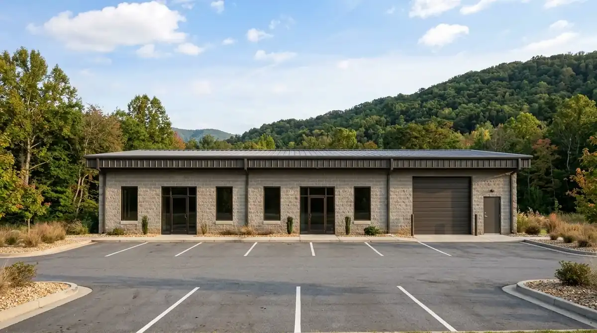 Single-story commercial flex building with split-face block walls, glass entry doors, a roll-up loading door, and an empty parking apron, with wooded hills behind it